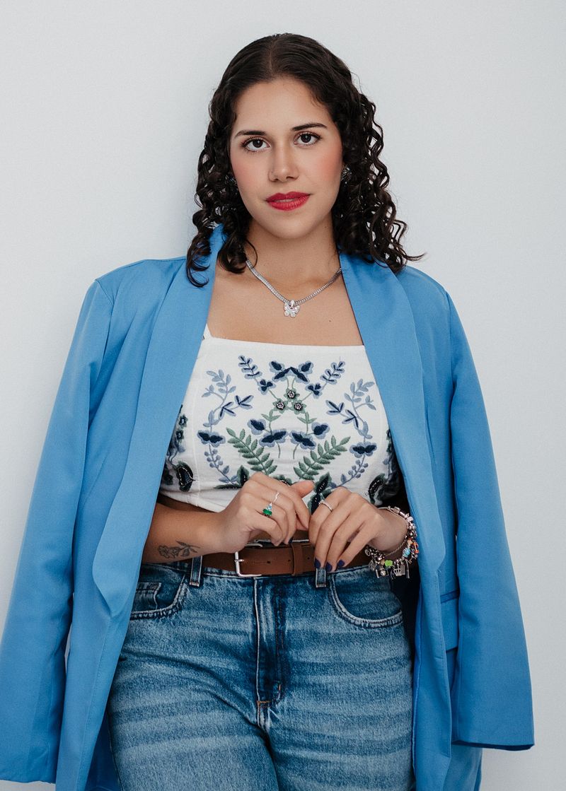 Nicoli Nascimento no consultório, de blazer azul e blusa bordada, encostada na parede